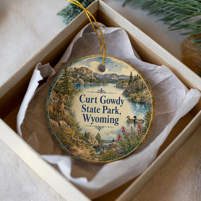 A decorative ornament in a box, featuring a scenic landscape with mountains, a lake, and wildlife, including a goat and a duck.