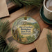 A round ornament with a painting of a cabin and a rifle, surrounded by pine branches and a cup of coffee on a wooden surface.