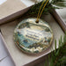 A decorative ornament in a box, featuring a scenic landscape with a lighthouse, water, and greenery.