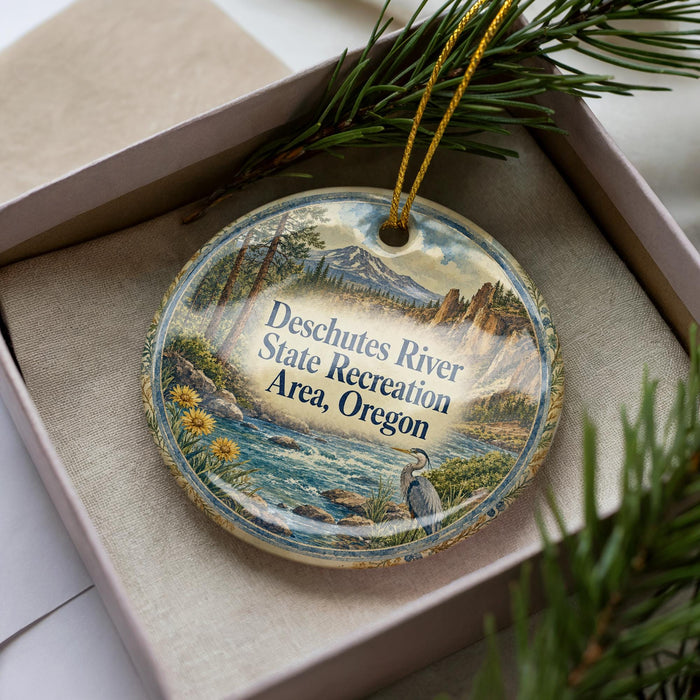 A decorative ornament with a scenic landscape of a river, mountains, and pine trees, placed in a box with a pine branch.