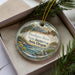 A decorative ornament with a scenic landscape of a river, mountains, and pine trees, placed in a box with a pine branch.