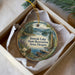 A decorative ornament in a box, featuring a scenic landscape with a lake, mountains, pine trees, and a boat on the water.