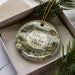 A decorative ornament in a box, featuring a scenic landscape with a waterfall and pine trees.