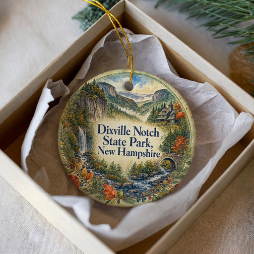 A decorative ornament in a box, featuring a scenic landscape with mountains, a waterfall, and a bridge.