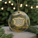 A decorative ornament featuring a scenic landscape with a monument, trees, and a cannon, hanging in front of a Christmas tree with lights.