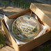 A decorative ornament in a brown box, featuring a scenic landscape with mountains, water, and greenery.