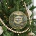 A round ornament hanging on a Christmas tree, featuring a scenic landscape with a river, a bridge, and a bird.