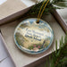 A decorative ornament with a beach scene and text, placed in a box with pine branches.
