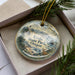 A decorative ornament with a landscape scene featuring mountains, pine trees, and a lighthouse, placed inside a box with a pine branch.
