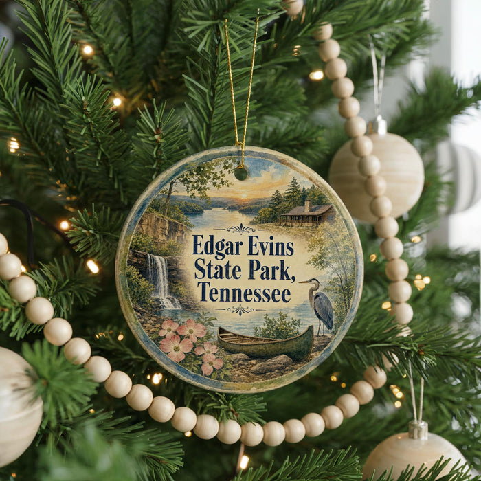 A round ornament hanging on a Christmas tree, featuring a scenic landscape with a waterfall, a bird, and a canoe.
