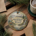 A decorative ornament featuring a scenic landscape with a waterfall, pine trees, and mountains, hanging on a wooden surface.