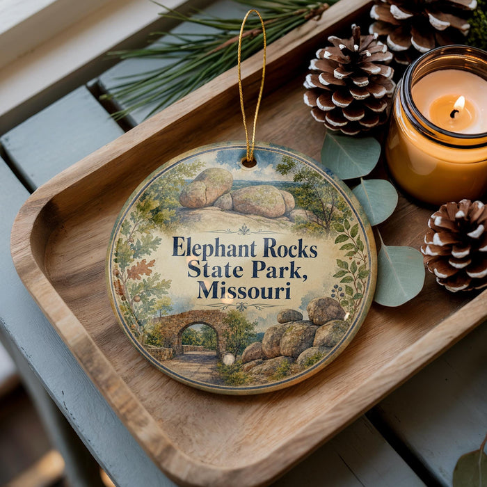 A decorative ornament with a scenic landscape of Elephant Rocks State Park, Missouri, featuring a bridge and boulders, is displayed on a wooden tray alongside pine cones and a lit candle.
