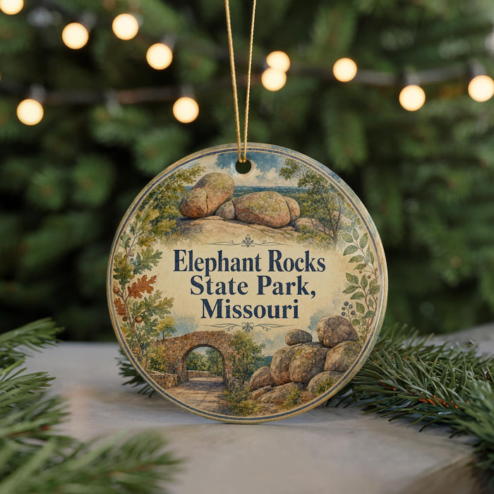 A round ornament with a scenic illustration of Elephant Rocks State Park, Missouri, featuring a bridge and boulders, is displayed on a table with pine branches and twinkling lights in the background.