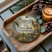 A wooden tray with a decorative ornament featuring a scenic image of a deer in a forest, along with pine cones and a lit candle.