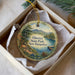 A decorative ornament in a box, featuring a scenic landscape with a lake, mountains, and pine trees.