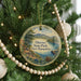 A round ornament hanging on a Christmas tree, featuring a scenic landscape with mountains, trees, and a lake.