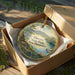 A decorative ornament in a wooden box, featuring a scenic landscape with a lake, mountains, and pine trees.