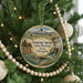 A round ornament hanging on a Christmas tree, featuring a scenic landscape with mountains, a river, and a wagon.