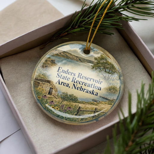 A decorative ornament in a box, featuring a scenic landscape with a body of water, trees, and a small building.