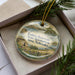 A decorative ornament in a box, featuring a scenic landscape with a body of water, trees, and a small building.