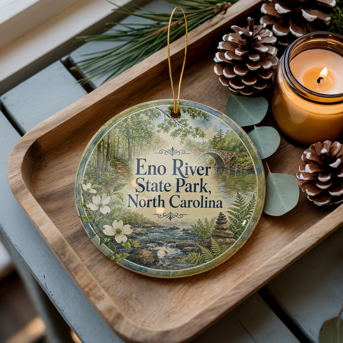 A wooden tray with a decorative ornament featuring a scenic image of Eno River State Park, North Carolina, along with pine cones and a lit candle.