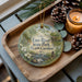 A wooden tray with a decorative ornament featuring a scenic image of Eno River State Park, North Carolina, along with pine cones and a lit candle.