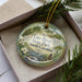 A decorative ornament in a box, featuring a scenic landscape with a river, trees, and flowers.