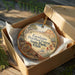 A round ornament in a box, featuring a scenic landscape with mountains, trees, and a winding path, with the text "Escalante Petrified Forest State Park, Utah" printed on it.
