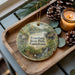 A decorative ornament featuring a scenic image of Evansburg State Park, Pennsylvania, with pine cones and a lit candle on a wooden tray.