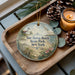 A decorative ornament with a lighthouse and beach scene, placed on a wooden tray next to pine cones and a lit candle.