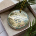 A decorative ornament in a box, featuring a lighthouse and a beach scene, with the text "Fair Haven State Park, New York" on it.
