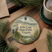 A round ornament with a scenic landscape of Cape State Park, Virginia, featuring a bird and a path, is displayed on a wooden surface alongside a cup of coffee and some pine branches.