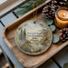 A wooden tray with a decorative ornament featuring a scenic landscape, pine cones, and a lit candle.