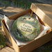 A round ornament in a box, featuring a scenic landscape with a building and a river, with the text "Farny State Park New Jersey" printed on it.