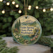 A round ornament with a scenic landscape of a mountain, river, and cabin, surrounded by pine trees and flowers, hangs in front of a blurred Christmas tree with lights.