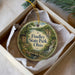 A round ornament with a scenic landscape of Findley State Park, Ohio, displayed in a box with a white tissue paper lining.