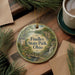 A decorative ornament with a scenic landscape of Findley State Park, Ohio, featuring a waterfall and a bridge, is displayed on a wooden surface alongside a cup of coffee and some pine branches.