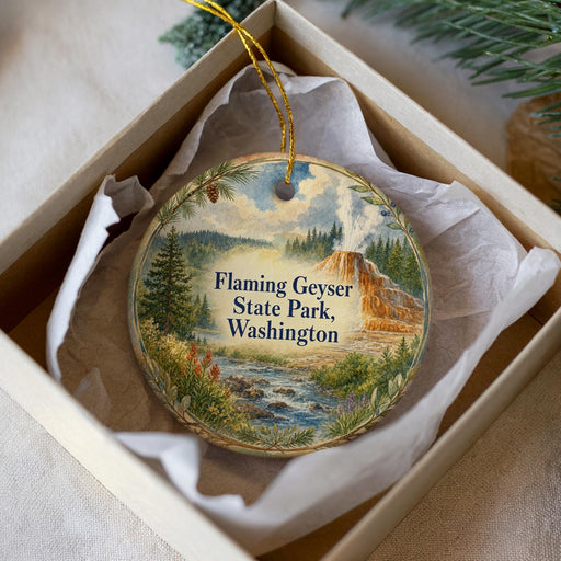 A decorative ornament in a box, featuring a scenic landscape with a mountain, river, and pine trees.