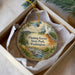 A decorative ornament in a box, featuring a scenic landscape with a mountain, river, and pine trees.