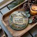 A decorative ornament featuring a lighthouse and a scenic landscape, placed on a wooden tray alongside pine cones and a lit candle.