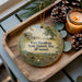 A decorative ornament with a picture of Fort Davidson State Historic Site in Missouri, featuring a building, trees, and a flag, placed on a wooden tray alongside pine cones and a lit candle.