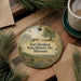A decorative ornament with a painting of Fort Davidson State Historic Site in Missouri, featuring a building, trees, and a flag, is displayed on a wooden surface alongside a cup of coffee and some papers.