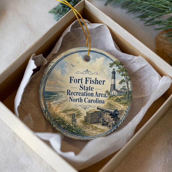 A round ornament with a lighthouse and beach scene, placed in a box with a white tissue paper lining.