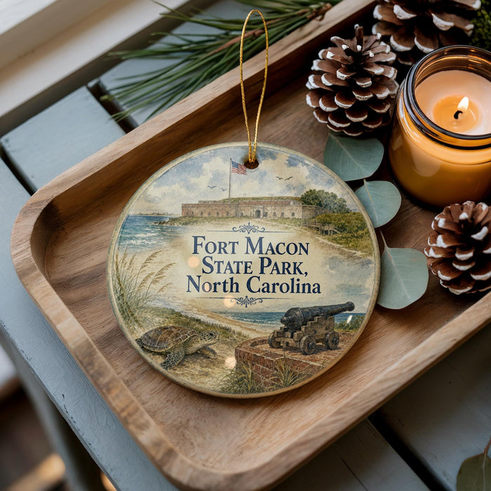 A decorative ornament featuring a scene from Fort Macon State Park, North Carolina, with a cannon and a turtle, placed on a wooden tray alongside pine cones and a lit candle.