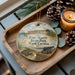 A decorative ornament featuring a scene from Fort Macon State Park, North Carolina, with a cannon and a turtle, placed on a wooden tray alongside pine cones and a lit candle.