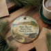 A round ornament with a picture of Fort Macon State Park, North Carolina, featuring a turtle, a cannon, and a building, is displayed on a wooden surface alongside a cup of coffee and some pine branches.