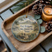 A decorative ornament with a picture of Fort McKayett, the historic site in Texas, sits on a wooden tray alongside pine cones and a lit candle.