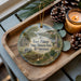 A decorative ornament with a scenic image of Fort Pillow State Historic Park, Tennessee, is displayed on a wooden tray alongside pine cones and a lit candle.