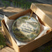 A decorative ornament in a wooden box, featuring a scenic landscape with a house, trees, and a body of water.