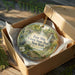 A decorative ornament in a brown box, featuring a scenic landscape with a river, trees, and wildlife.